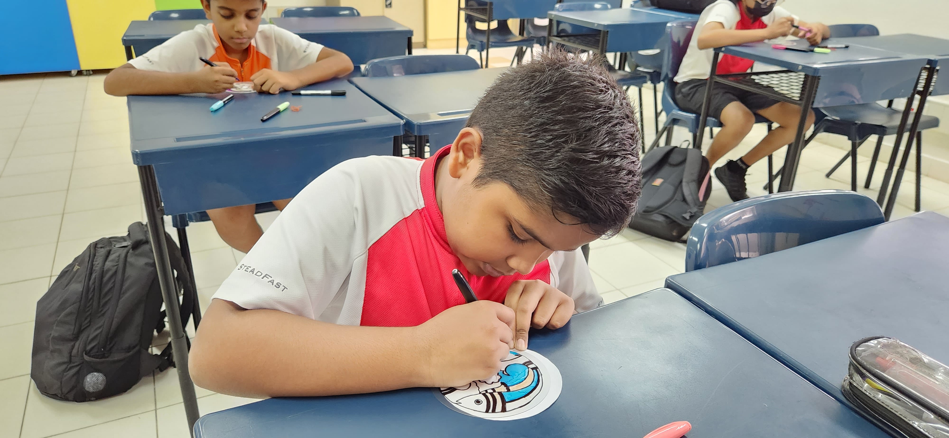 A boy in a white and red shirt colors a circular drawing at a blue desk in a classroom with other students.